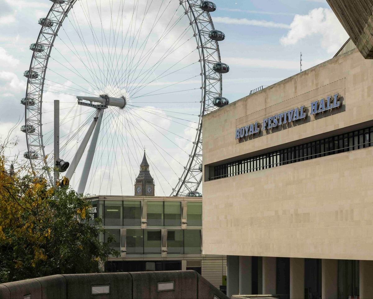 London Eye and Royal Festival Hall in London, UK, with the large observation wheel rising behind the beige stone building under a cloudy sky.