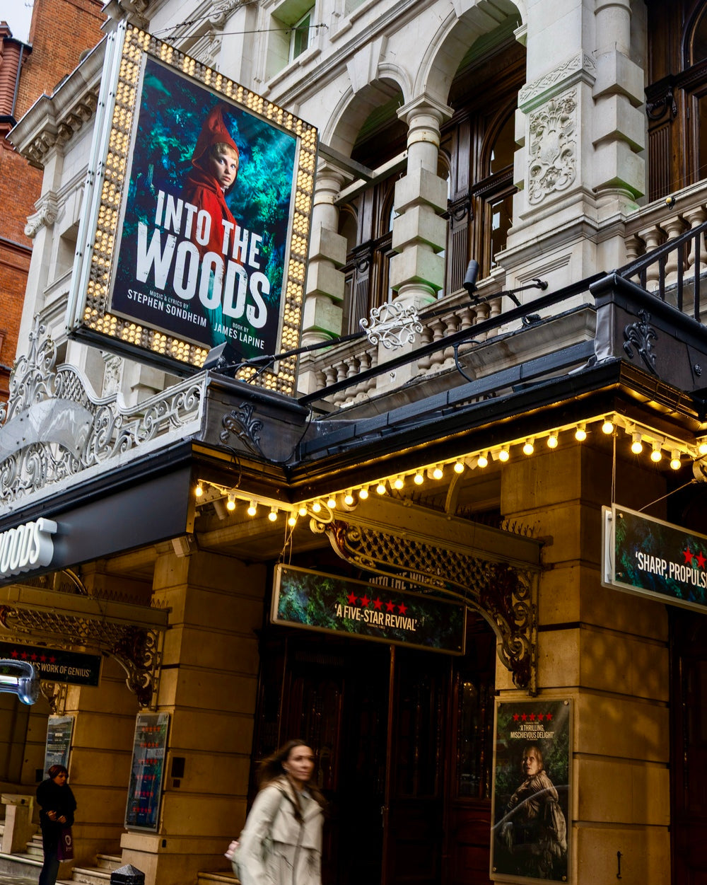 A real outdoor street scene in front of a theater building, with a large marquee reading 'Into the Woods' and a woman walking in the foreground. Because a real environment and a person are present, this is Lifestyle rather than Product/Packaging.